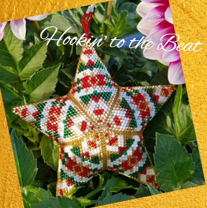 May include: A star-shaped ornament made of beads in red, white, green, and gold colors. The ornament is hanging from a green plant with white flowers. The text "Hookin' to the Beat" is visible in the image.