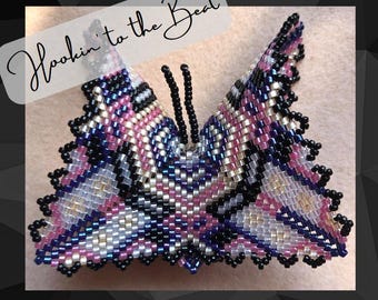 Könnte beinhalten: Eine Brosche in Form eines Schmetterlings aus Perlen mit einem geometrischen Muster in Rosa, Weiß und Blau. Der Schmetterling ist aus kleinen, bunten Perlen gefertigt und hat einen schwarzen Umriss.