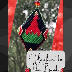 May include: A red, green, black, and gold beaded ornament shaped like a diamond with a bell hanging from the bottom. The ornament has the text "Hookin' to the Beat" on a white background.