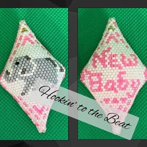 Op de afbeelding: Ruitvormige, met kralen bezette hanger met een baby-olifantontwerp in grijs en wit, geaccentueerd met roze. De achterkant toont de woorden "NEW BABY" in roze. De hanger staat tegen een groene achtergrond.