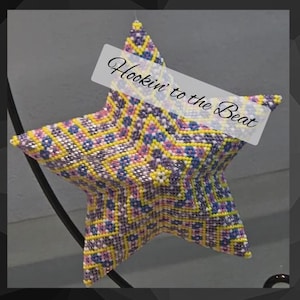 May include: A beaded star ornament with a geometric pattern in yellow, purple, blue, and white. The star has a white banner across it with the text "Hookin' to the Beat". The ornament is hanging from a black metal stand.