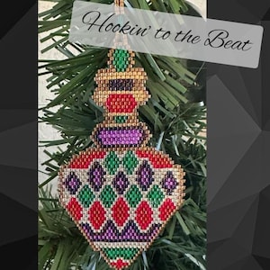 May include: A colorful beaded Christmas ornament in the shape of a teardrop with a gold, red, green, and purple pattern. The ornament is hanging on a green Christmas tree branch. The text "Hookin' to the Beat" is written in white on a black background.