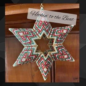 Può includere: Un ornamento a forma di stella in perline rosse, verdi e dorate con un motivo geometrico. L'ornamento è appeso a una superficie di legno e ha il testo "Hookin' to the Beat" sopra.