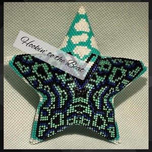 Pode incluir: Um ornamento em forma de estrela de cinco pontas feito de pequenas contas em tons de azul-petróleo, azul escuro, preto e branco. A ponta superior apresenta um padrão branco e azul-petróleo, enquanto as seções inferiores exibem um design geométrico azul escuro e preto. O texto Hookin' to the Beat está impresso na diagonal no centro.