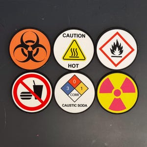 May include: Six round warning signs with different symbols and text. The signs are white with black borders and feature various symbols, including a biohazard symbol, a fire symbol, a radiation symbol, a hot symbol, a no food or drink symbol, and a corrosive symbol. The text on the signs includes "CAUTION HOT", "CAUSTIC SODA", and "CORR".