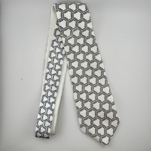 May include: A white necktie with a repeating black geometric honeycomb pattern. The tie is made of a smooth fabric and is displayed on a white background. The design is consistent across the entire tie.
