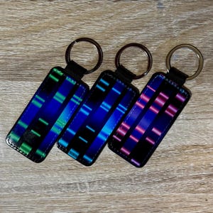 May include: Three rectangular keychains with a black leather base and a metal keyring. Each keychain features a unique design with a gradient of colors: green and blue, blue, and pink and purple. The keychains are displayed on a wooden surface.