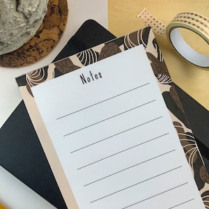 May include: A white notepad with a brown and beige abstract pattern border. The notepad has lined pages and the text "Notes" at the top. The bottom of the notepad has the text "~be kind to yourself~" in black.