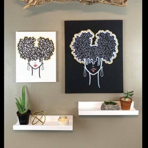 Puede incluir: Dos cuadros enmarcados de una mujer con un peinado afro. Los cuadros est&aacute;n en estantes blancos con una planta en maceta, una vela y una esfera decorativa. Un trozo de madera a la deriva cuelga del techo sobre los cuadros.