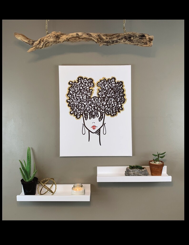 Puede incluir: Un dibujo en blanco y negro de una mujer con un gran peinado afro. El afro est&aacute; contorneado con purpurina dorada. El dibujo est&aacute; en un lienzo blanco y est&aacute; colgado en una pared con una rama de madera encima. Hay dos estantes blancos debajo del lienzo, uno con una vela y una esfera dorada, y el otro con una planta en maceta y una peque&ntilde;a roca.
