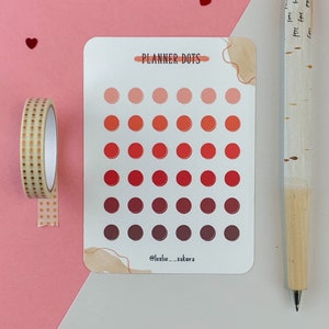 May include: A sheet of planner dots stickers with a gradient of red, orange, and pink colors. The sheet is white with a brown watercolor design in the corner. The text "PLANNER DOTS" is printed at the top of the sheet. The text "@leslie__sakura" is printed at the bottom of the sheet.