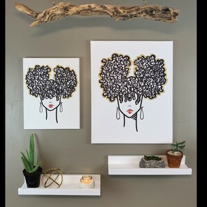 Puede incluir: Dos impresiones en lienzo en blanco y negro de una mujer con un peinado afro. Las impresiones est&aacute;n colgadas en una pared con una rama de madera flotante encima. Hay dos estantes blancos debajo de las impresiones, uno con una vela y una esfera decorativa, y el otro con una peque&ntilde;a planta en maceta y una roca.
