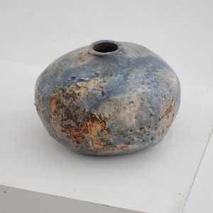 Könnte beinhalten: Eine kleine, runde Keramikvase mit einer texturierten, unebenen Oberfläche. Die Glasur zeigt eine gesprenkelte Mischung aus staubigen Blau-, Grau- und erdigen Brauntönen, mit einer kleinen kreisförmigen Öffnung oben und einer bauchigen Form.