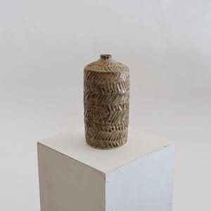 Op de afbeelding: Een cilindrische keramische vaas met een textuurrijk, uitgesneden visgraatpatroon over het oppervlak. Het object heeft een genuanceerd glazuur in aardse tinten bruin, beige en gedempt groen, afgewerkt met een smalle hals en een kleine opening.