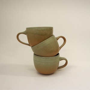 Peut inclure: Trois tasses en céramique empilées dans des tons de vert et de brun. Les tasses ont une forme arrondie et une surface légèrement texturée.
