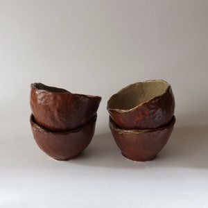 Puede incluir: Cuatro cuencos de cerámica marrón apilados en dos juegos de dos. Los cuencos tienen una superficie rugosa y texturizada y una forma ligeramente irregular.