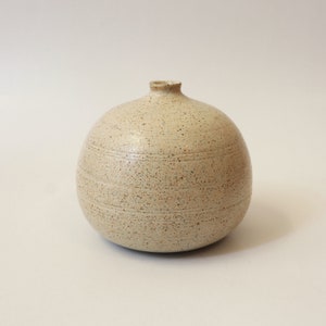 May include: A small, round, speckled ceramic vase with a narrow opening. The vase is a light beige color with a subtle, textured finish.
