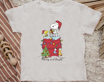 May include: White t-shirt featuring a cartoon Snoopy and Woodstock in Santa hats, atop a red doghouse decorated with Christmas lights. A small Christmas tree, stocking, and the words "Merry and bright" are also visible.