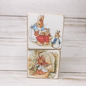 May include: Two wooden blocks with illustrations of Peter Rabbit. The top block shows Peter Rabbit with two other rabbits. The bottom block shows Peter Rabbit walking in the woods.