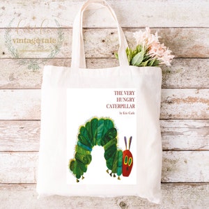 May include: A white canvas tote bag with a colorful illustration of The Very Hungry Caterpillar by Eric Carle. The text "THE VERY HUNGRY CATERPILLAR by Eric Carle" is printed in brown on the bag.