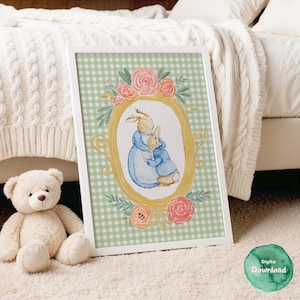 May include: A framed watercolour print depicting two rabbits embracing, set within a gold oval frame decorated with pink roses and foliage. The background is a green and white gingham pattern. A teddy bear sits in front of the framed print.