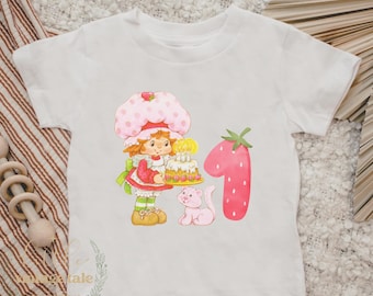 May include: White t-shirt with a graphic of Strawberry Shortcake holding a birthday cake and a pink cat. The number 1 is in the shape of a strawberry.