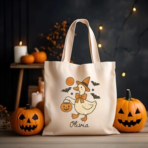 May include: Beige Halloween tote bag featuring a cartoon duck wearing a witch hat and holding a jack-o'-lantern. The design includes bats and a full moon. The name "Olivia" is printed below the duck. Two carved pumpkins and candles are in the background.