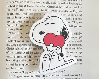 May include: A white and black cartoon dog holding a red heart. The dog is sitting down with its legs crossed. The dog has a black nose and eyes. The heart is red and has a white outline.
