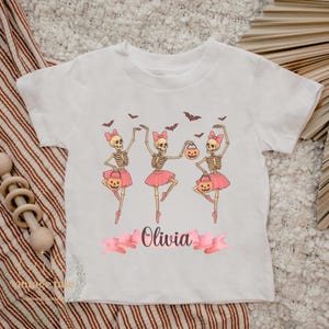 Puede incluir: Camiseta blanca con diseño de Halloween. Muestra tres bailarinas esqueleto con tutús rosas, sosteniendo calabazas y lazos rosas. El nombre "Olivia" está escrito en cursiva sobre una cinta rosa.