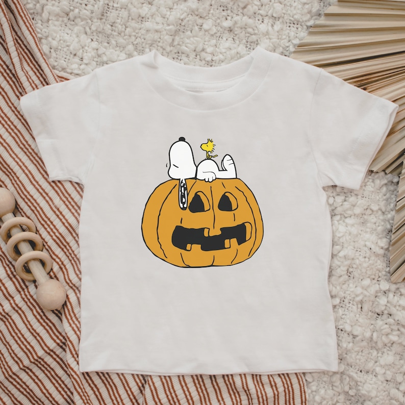 May include: White t-shirt with a cartoon image of Snoopy and Woodstock on a jack-o'-lantern.