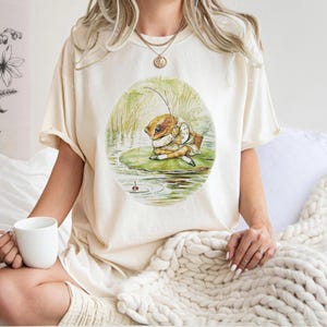 May include: Cream-colored t-shirt featuring a whimsical illustration of a frog in a coat and hat, sitting on a lily pad and fishing. The frog is depicted in a vintage, storybook style. The shirt is a relaxed fit.