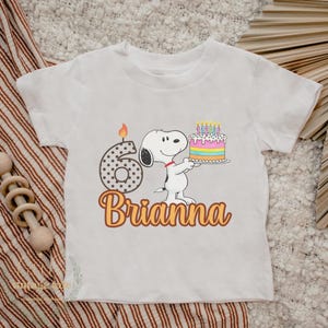 May include: White toddler t-shirt featuring a cartoon Snoopy character holding a birthday cake with candles. The number 6 with a lit candle and the name "Brianna" are also displayed in a playful font.