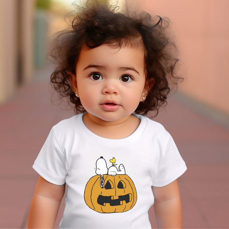 May include: A white t-shirt with a cartoon image of Snoopy and Woodstock sitting on top of a jack-o'-lantern. The child wearing the shirt has brown curly hair.