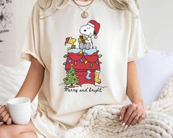 May include: Cream-colored t-shirt featuring a festive Snoopy graphic. Snoopy, wearing a Santa hat, is atop his doghouse, decorated with Christmas lights and a tree. The text "Merry and bright" is below the graphic.