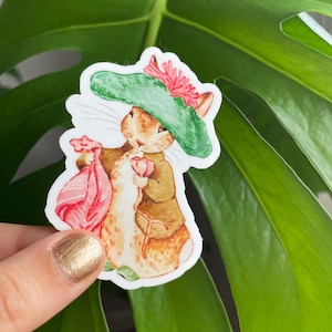 May include: A sticker featuring a cartoon rabbit wearing a green hat with a red flower. The rabbit is holding a pink flower and a pink and white striped bag.