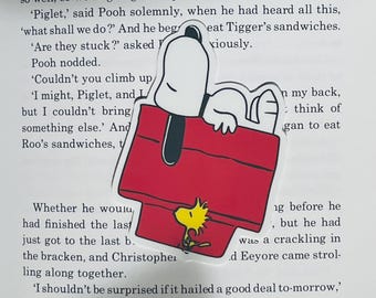 May include: A die-cut sticker featuring Snoopy, the cartoon dog, lying on top of his red doghouse. A yellow Woodstock is perched on the doghouse. The sticker is placed on a book page.