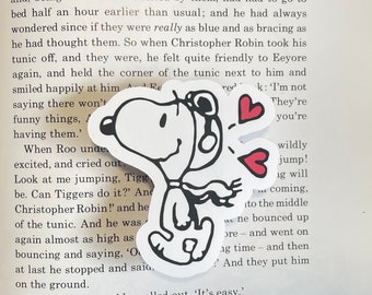 May include: A black and white cartoon sticker of Snoopy wearing a pilot's hat and scarf, with two red hearts above him. The sticker is on a page of a book with text that reads "I'm not They're you're wildly jump! ig will coming, to the middle av ne bounced up ore - and went on ng time - and then ? And they put him"