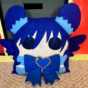 May include: Blue and white plush pillow with a heart-shaped bow and wings. The pillow has a cartoon face with large eyes and blue hair.