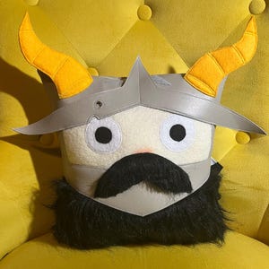 May include: A square decorative pillow with a Viking helmet design. The pillow features a light gray helmet with yellow horns, a white face, black eyes, a black mustache, and a black beard. The pillow is on a yellow chair.