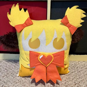 May include: A yellow plush pillow with a heart-shaped bow and two yellow pigtails. The pillow has a white face with brown eyes and a red mouth.