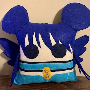 May include: A handmade, square pillow featuring a cartoon character design. The pillow has a white face with large black eyes, blue hair, and blue wings. The character wears a blue and white outfit with a gold accent. The pillow is made of felt.