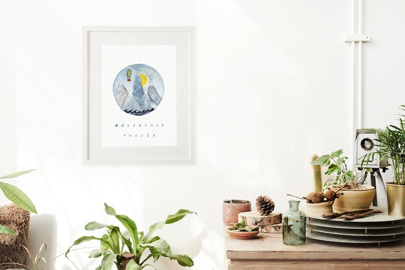 K&ouml;nnte beinhalten: Ein wei&szlig;er gerahmter Druck mit einer Aquarell-Illustration von Bergen, einem Hei&szlig;luftballon und einer Sonne. Der Text "adventure awaits" steht unter der Illustration.