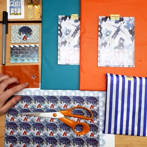 K&ouml;nnte beinhalten: Eine flache Anordnung von Bastelmaterialien zum Verpacken von Geschenken. Die Materialien umfassen eine Schere, Geschenkpapier mit einem Berg- und Zeltmuster, ein blaues und wei&szlig; gestreiftes Stoffst&uuml;ck und zwei Karten mit dem Text "Unterst&uuml;tze die Macher".