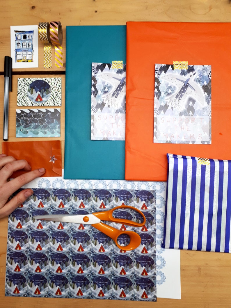 K&ouml;nnte beinhalten: Eine flache Anordnung von Bastelmaterialien zum Verpacken von Geschenken. Die Materialien umfassen eine Schere, Geschenkpapier mit einem Berg- und Zeltmuster, ein blaues und wei&szlig; gestreiftes Stoffst&uuml;ck und zwei Karten mit dem Text "Unterst&uuml;tze die Macher".
