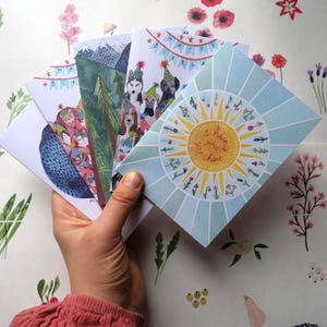 May include: A hand holds a collection of illustrated greeting cards. The cards feature various designs, including a mountain scene, dogs wearing party hats, and a sun with the text "sending you sunshine". The background is a floral pattern.