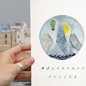 K&ouml;nnte beinhalten: Eine Aquarell-Illustration eines Hei&szlig;luftballons, der &uuml;ber Berge mit Sonne und Wellen fliegt. Der Text "adventure awaits" steht unter der Illustration.