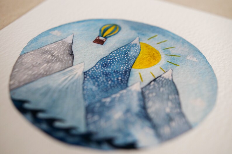 K&ouml;nnte beinhalten: Ein Aquarellbild eines blauen Himmels mit einer gelben Sonne und einem Hei&szlig;luftballon. Die Berge sind in Blaut&ouml;nen und Grau gemalt.