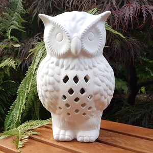 May include: White ceramic owl lantern with a cut-out design. The owl is standing on a wooden table.