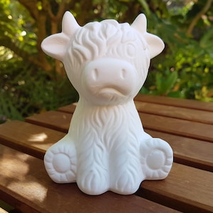 May include: A white ceramic Highland cow ornament with detailed fur, horns, and a gentle expression. The cow is sitting on a wooden surface, with a blurred green background.