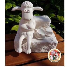 May include: A white ceramic figurine of Shaun the Sheep sitting on a bench with a cup of tea and a slice of cake. The figurine is officially licensed and features the characters Shaun the Sheep, Bitzer, and Timmy.